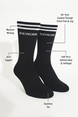 Two black BODYBORN crew-length socks with white stripe detailing, displayed upright and labeled with features including moisture-wicking fabric, arch support, all-over cushioning through the foot and leg, antimicrobial and antifungal half-terry construction and seamless toe
