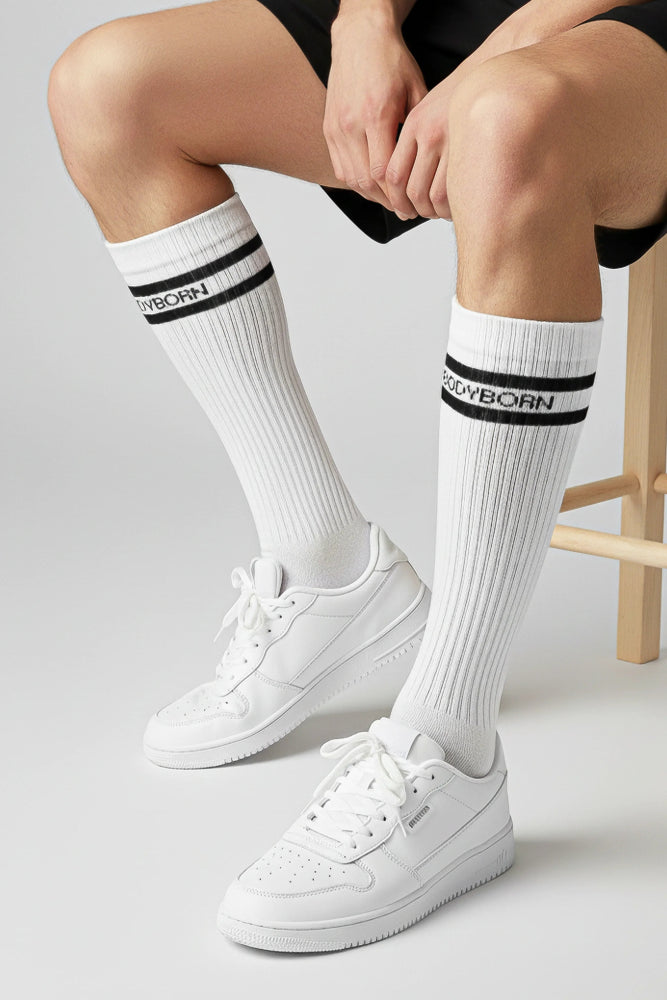 White socks with black stripes and 'BODYBORN' branding worn by a woman sitting on a stool in crew size