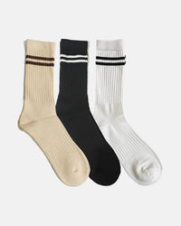 Three crew-length socks arranged side by side one beige with brown stripes, one black with white stripes, and one white with black stripes each designed with rib-knit texture and two-stripe detailing