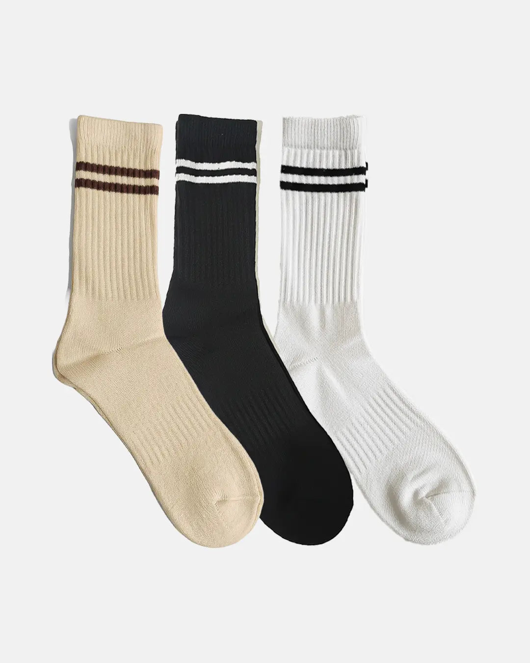 Three crew-length socks arranged side by side one beige with brown stripes, one black with white stripes, and one white with black stripes each designed with rib-knit texture and two-stripe detailing
